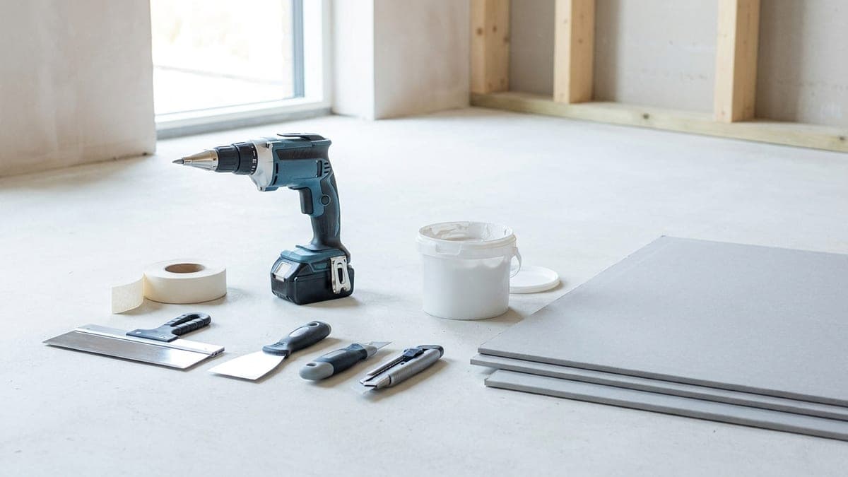 Outils de plaquiste posés devant des plaques de plâtre : visseuse à placo, couteau à enduire, cutter et bande à joint